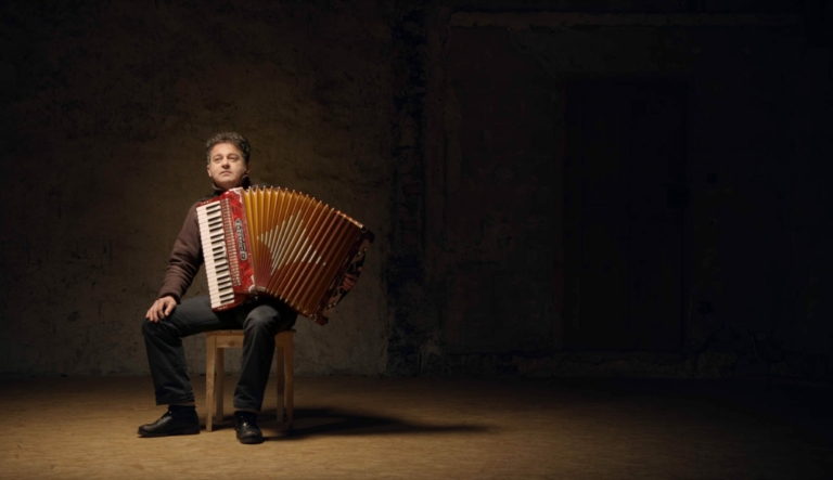 Eine Szene aus dem Film über Werner Pirchner: Wir sehen den Musiker Siggi Haider in einem sehr düsteren Raum mit einer Ziehharmonika auf einem Stuhl sitzen. Er sieht nachdenklich aus.