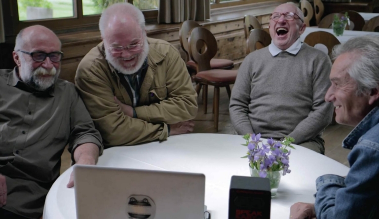 Eine Szene aus dem Film über Werner Pirchner: Wir sehen eine Runde älterer Männer, die gemeinsam rund um einen Tisch sitzen und lachend auf den Bildschirm eines Computers schauen.