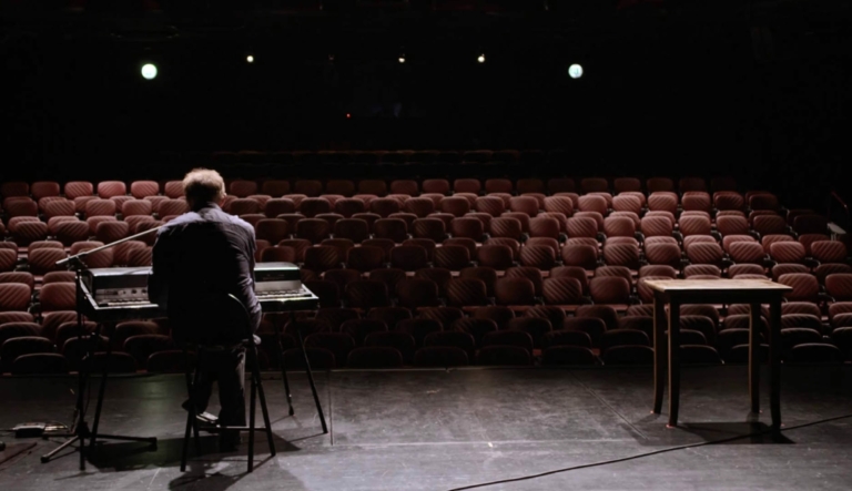 Eine Szene aus dem Film über Werner Pirchner: Wir sehen Josef Hader auf einer Bühne. Er sitzt mit dem Rücken zur Kamera an seinem Keyboard. Im Hintergrund serhen wir den leeren Zuschauerraum.