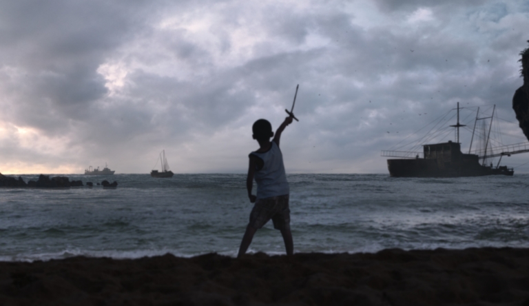 Nonoy steht mit einem Holzschwert am Strand und hält dieses kämpferisch den Schiffen entgegen.