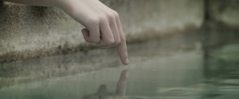 Wir sehen die Hand eines jungen Mädchens. Sie berührt mit dem Finger die spiegelglatte Wasseroberfläche eines Brunnens.